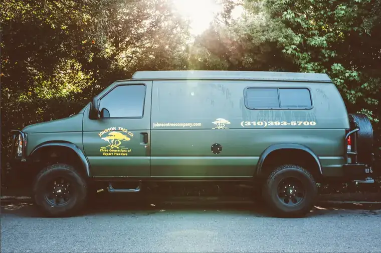 The Johnson Tree Company van, dark green with the yellow tree mark on the door, parked with the sun coming through the trees behind it.